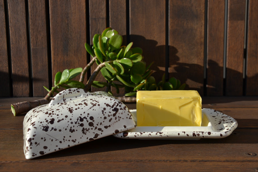 A speckled white ceramic butter dish with a matching lid and a rectangular yellow butter block, styled on a dark wooden surface with a green succulent plant in the background.