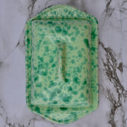 A green speckled ceramic butter dish with a matching lid, photographed from directly above on a white and gray marble surface.