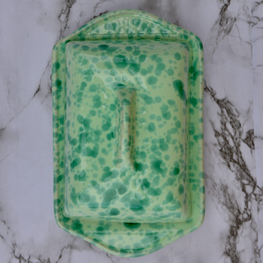 A green speckled ceramic butter dish with a matching lid, photographed from directly above on a white and gray marble surface.