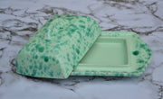 A green speckled ceramic butter dish with its lid propped open, set against a white and gray marble background.