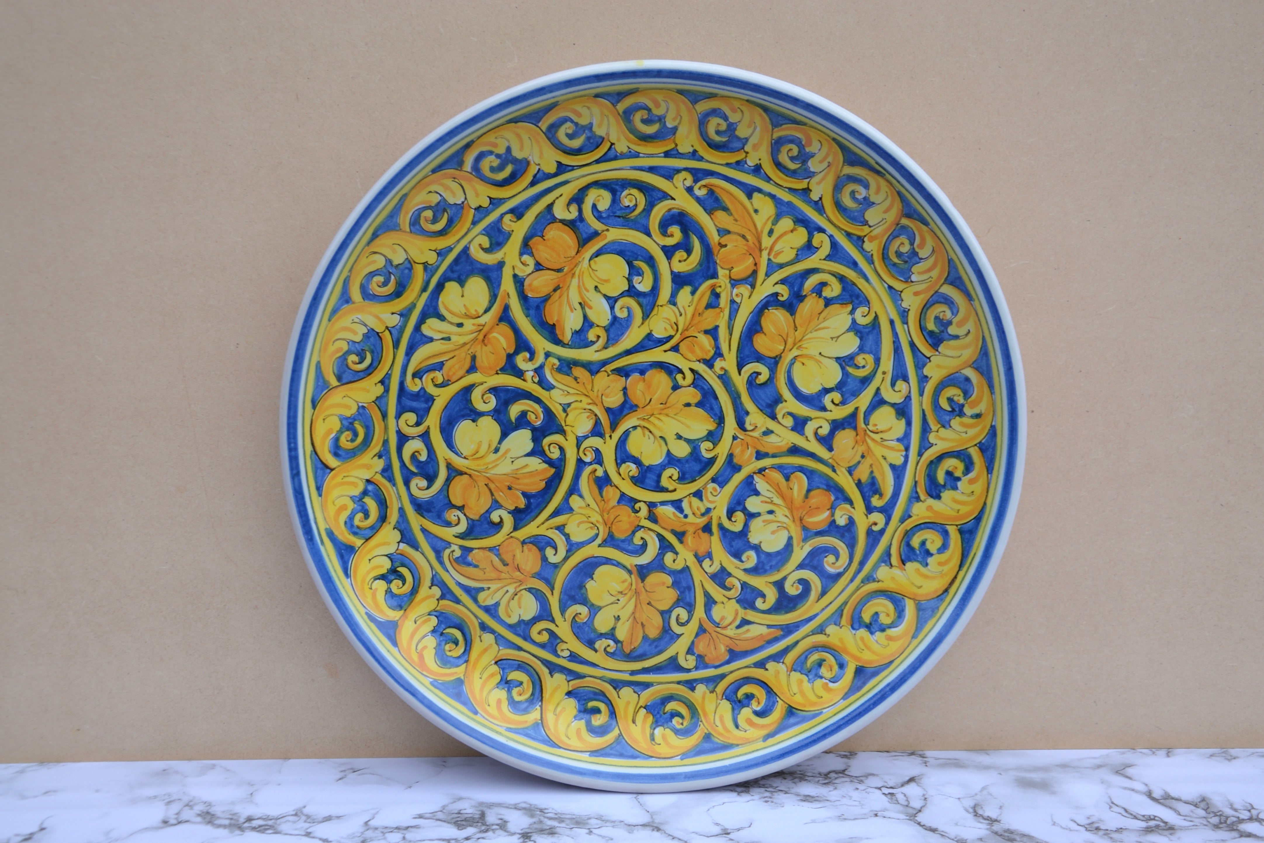 A round ceramic plate with a vibrant blue and yellow floral scroll pattern, displayed against a beige wall on a white marble surface.