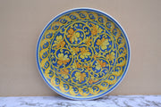 A round ceramic plate with a vibrant blue and yellow floral scroll pattern, displayed against a beige wall on a white marble surface.