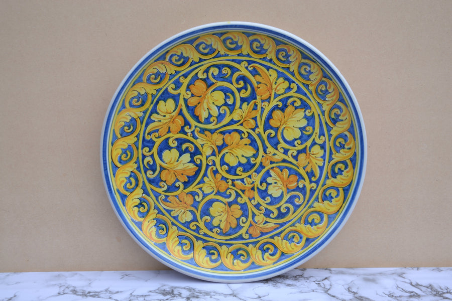 A round ceramic plate with a vibrant blue and yellow floral scroll pattern, displayed against a beige wall on a white marble surface.