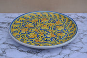 A round ceramic serving platter with a vibrant blue and yellow floral scroll pattern, displayed on a white and gray marble surface against a beige background.