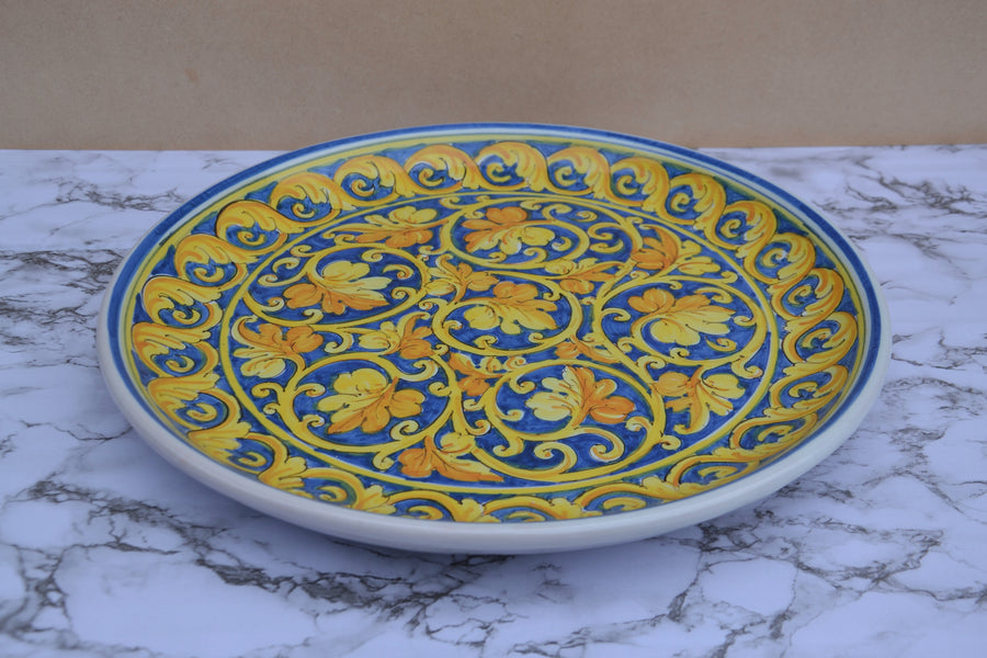 A round ceramic serving platter with a vibrant blue and yellow floral scroll pattern, displayed on a white and gray marble surface against a beige background.